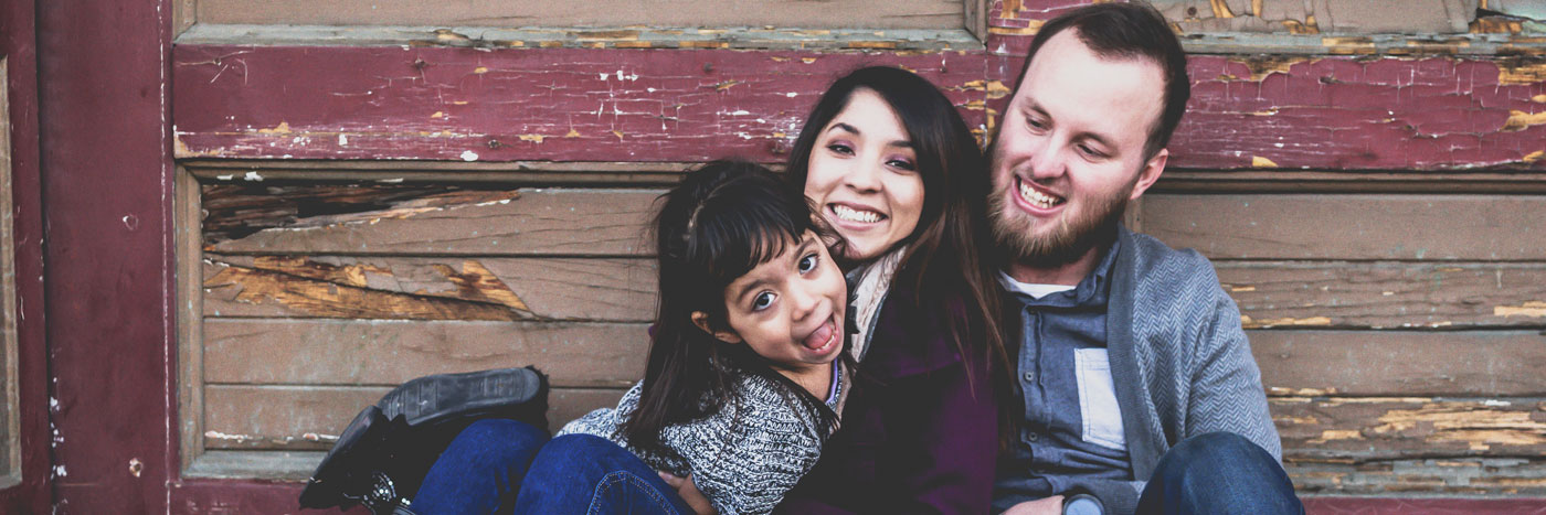 Mother, father, and daughter sitting against barn wall smiling